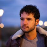 Portrait of a young man with curly hair wearing a leather jacket, captured outdoors at twilight.